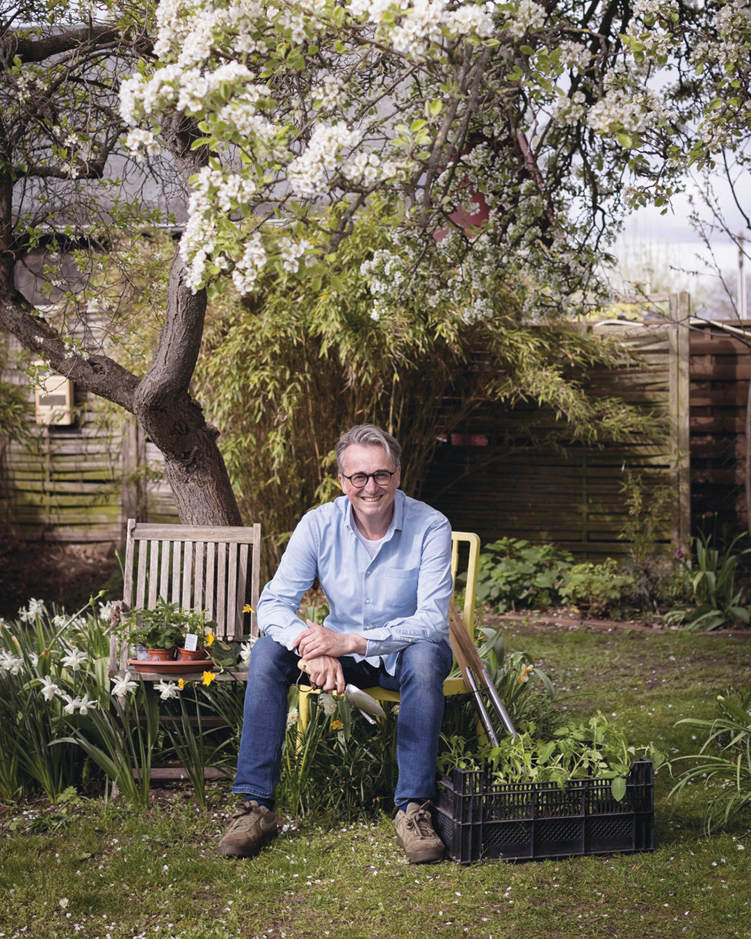 Horst Mager sitzt lächelnd auf einem Gartenstuhl unter einem blühenden Baum, umgeben von Pflanzen und Gartengeräten, und lädt ein zu seinem "Fr. 13.03.2026 | 13.00-16.00 HALBTAGESKURS Das Gartenjahr-Monat für Monat mit Horst Mager.
