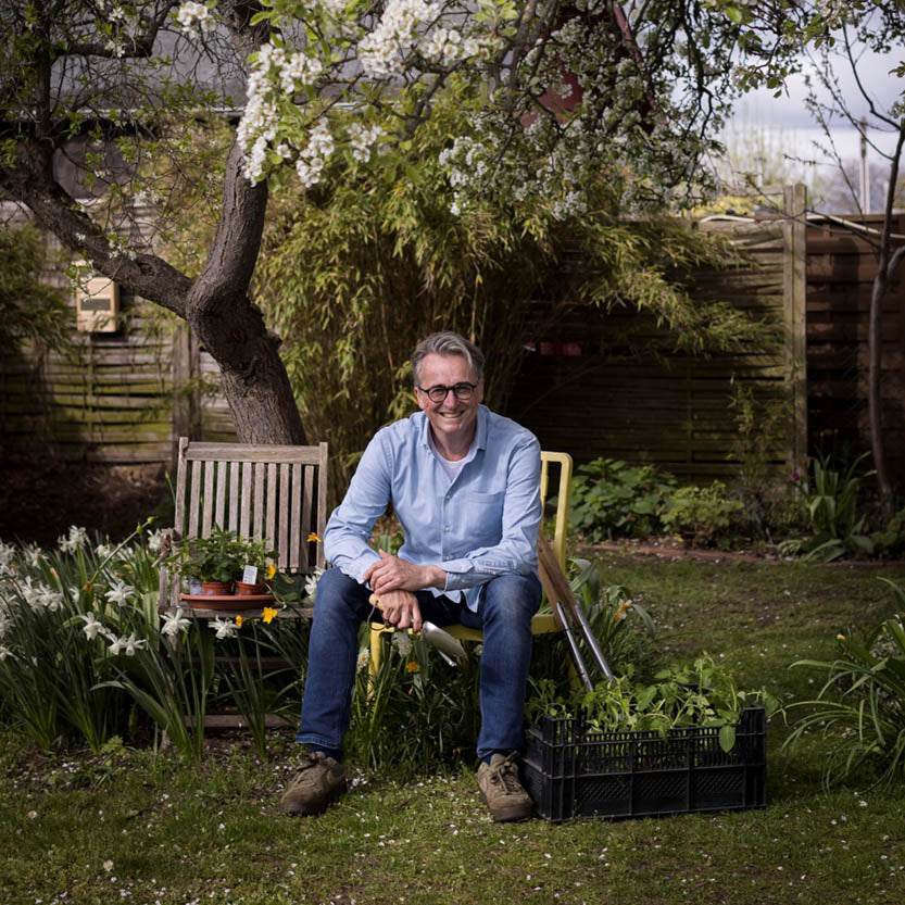 Ein Mann mit Brille und blauem Hemd sitzt zwischen Pflanzen, lächelt und ist bereit, Gartentipps zu geben. In der Nähe stehen Setzlinge und Werkzeuge für "Sa. 04.10.2025 | 13-16 Uhr HALBTAGESKURS - Gärtnern zu jeder Jahreszeit" mit Horst Mager, Königliche Gartenakademie.