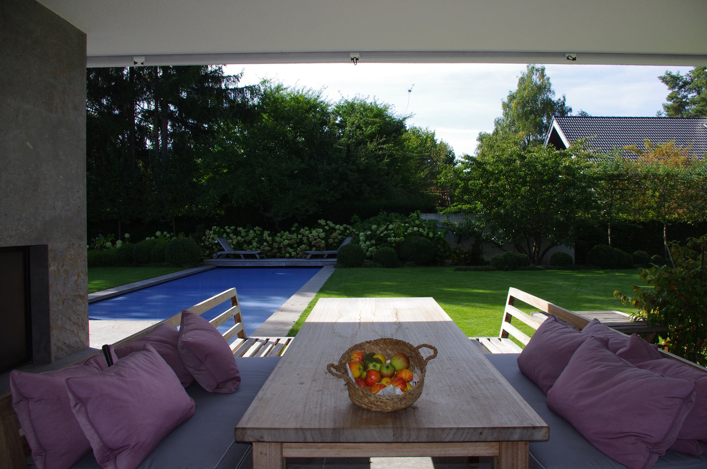 Unter einer überdachten Terrasse lädt ein hölzerner Esstisch mit Äpfeln zum Entspannen am Pool und im Garten ein und illustriert wunderschön Gabriella Papes "Mi. 18.02.2026 | 09.30-12.30 HALBTAGESKURS Der Garten als Lebensraum NEU.