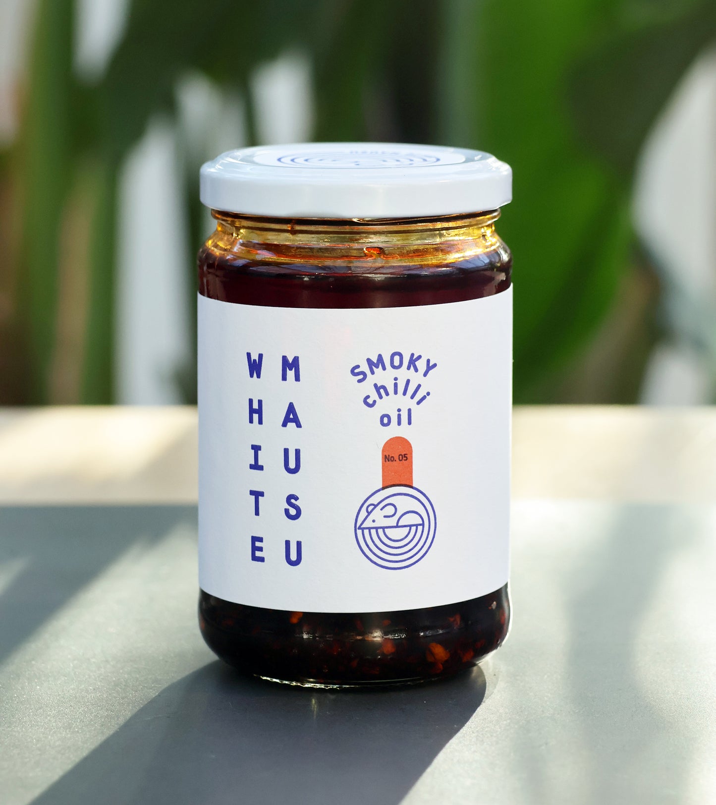Ein Glas White Mausu Ltd Rauchiges Chiliöl mit weißem Etikett und blauem Text steht auf einer grauen Fläche, grüne Pflanzen sind im Hintergrund leicht verschwommen.