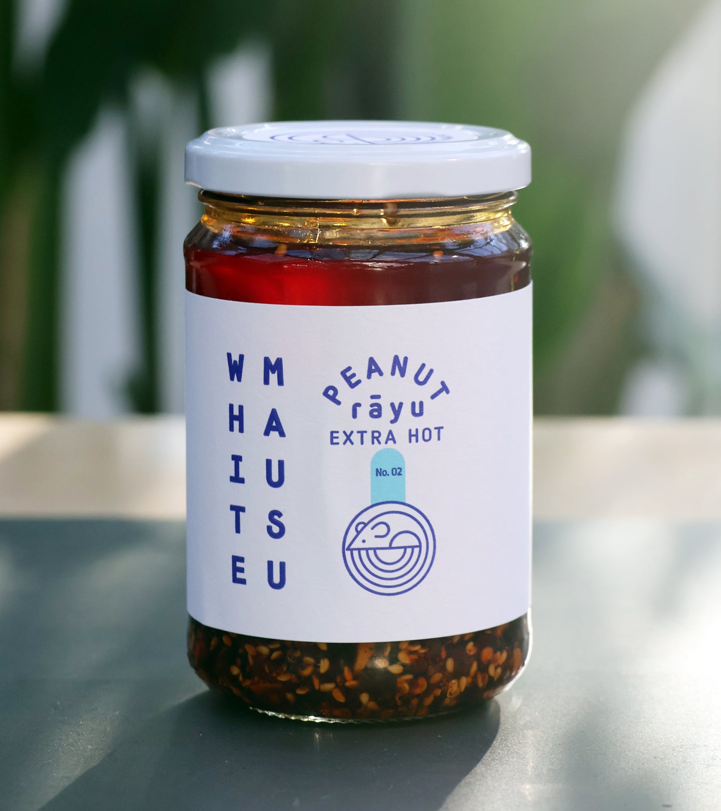 Ein Glasgefäß mit der Aufschrift Erdnuss Rayu Extra Hot von White Mausu Ltd steht auf einem Tisch. Es enthält extra scharfes Chili-Öl mit sichtbaren Samen und Stücken, verschlossen mit einem silbernen Deckel. Ein sanft verschwommener Hintergrund hebt den würzigen Inhalt hervor.