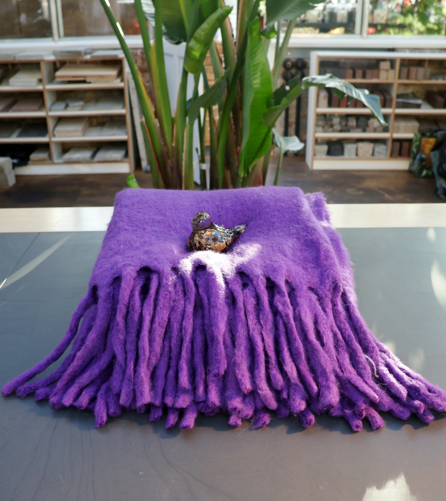 Die gefaltete Decke aus Wollmischung in Lila von Werner Voss liegt auf dem Tisch, dekoriert mit einer kleinen Vogel-Figur. Im Hintergrund befinden sich grüne Pflanzen und Regale mit verschiedenen Gegenständen.