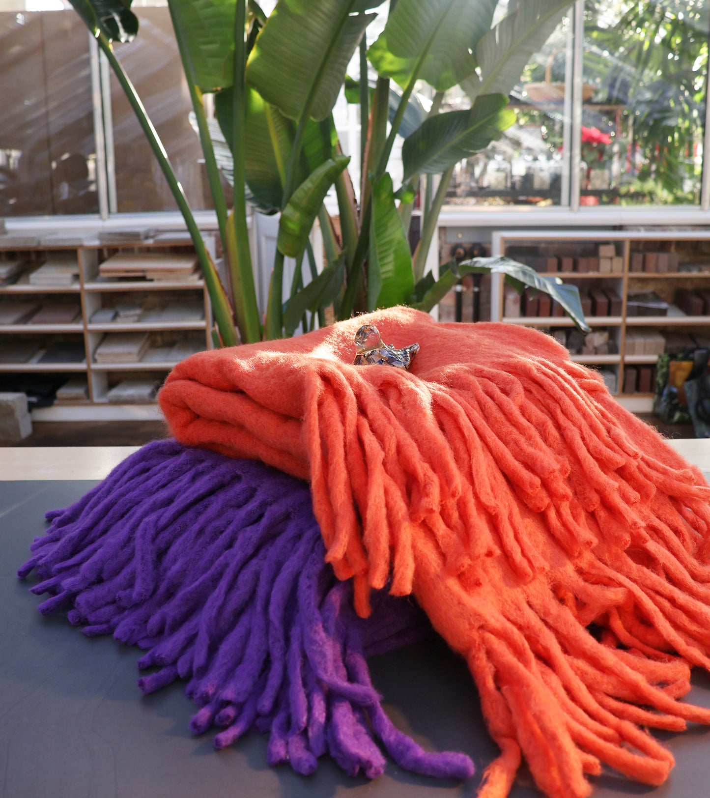 Eine dicke, gefranste Decke aus Wollmischung in Orange von Werner Voss liegt gefaltet auf einem Tisch unter einem kleinen Dekorationsobjekt. Sonnenbeschienenes Grün und Regale schaffen eine gemütliche, einladende Kulisse.