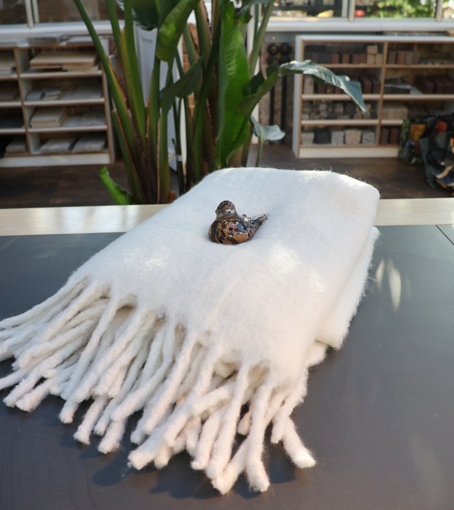 Die weiße Decke aus Wollmischung von Werner Voss ist auf einen Tisch gefaltet, ein kleiner Keramikvogel sitzt darauf; im Hintergrund sind Pflanzen sowie Regale mit Büchern und Materialien zu sehen - gemütliches Ambiente.
