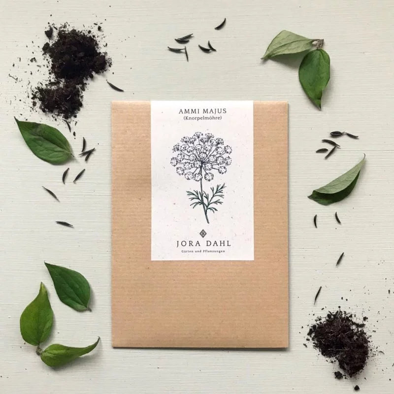 Das Jora Dahl | Ammi majus (Knorpelmöhre) Saatgutpaket ruht auf einer hellen Oberfläche mit verstreuter Erde, Samen und Blättern. Die Packung ist mit einer schwarzen, floralen Zeichnung und der Marke Jora Dahl versehen - perfekt für Ihren Schnittblumengarten.