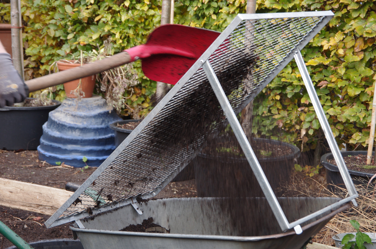 In Thea Carlins "WORKSHOP Kompost: Das wahre Gold des Gärtners" siebt eine Person mit einer roten Schaufel Erde und Kompost durch ein Netz in eine Schubkarre; im Hintergrund erscheinen Gartentöpfe, Grünzeug und Bodenaktivator.