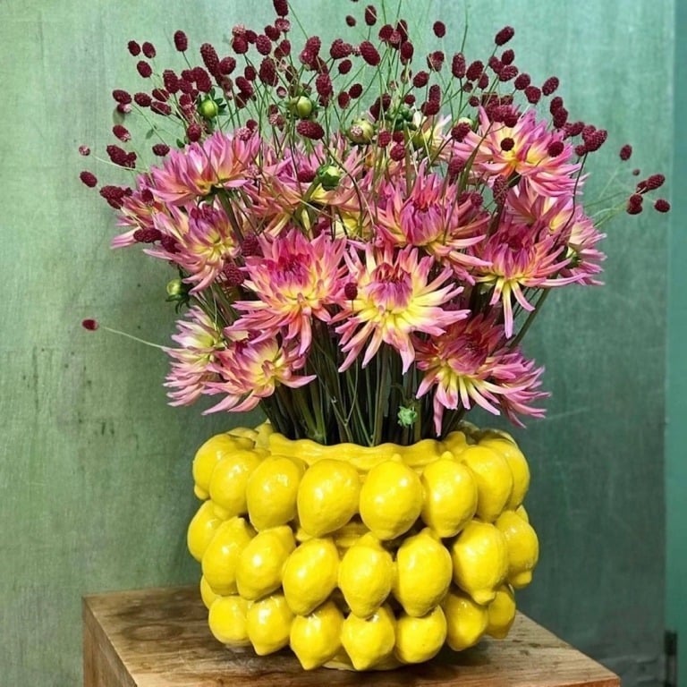 Ein Strauß aus rosa und gelben Blumen mit roten Knospen ist im Blumentopf von Des Pots | mit Zitronen in Gelb by Des Pots auf Holz vor grünem Hintergrund arrangiert und verleiht jedem Raum mediterrane Eleganz.