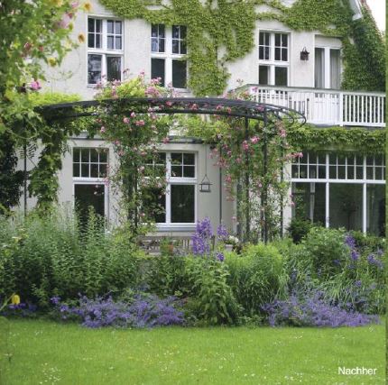 Ein üppiger Garten, gestaltet mit dem 5 Euro pro Quadratmeter Konzept – Gartendesign der Königlichen Gartenakademie, umgibt ein mit weißem Efeu bewachsenes Haus. Hohe Fenster, ein Balkon und eine Metalllaube mit Kletterrosen runden das elegante Bild ab.
