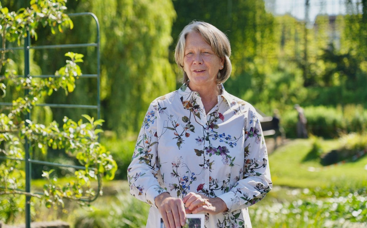 Eine Frau steht in einem sonnenbeschienenen Garten, hält ihr Smartphone in der Hand und trägt eine geblümte Bluse. Sie scheint sich für den "HALBTAGESKURS Grundlagen der Gartengestaltung" von Gabriella Pape an der Königlichen Gartenakademie am 06.11.2025 zu interessieren.
