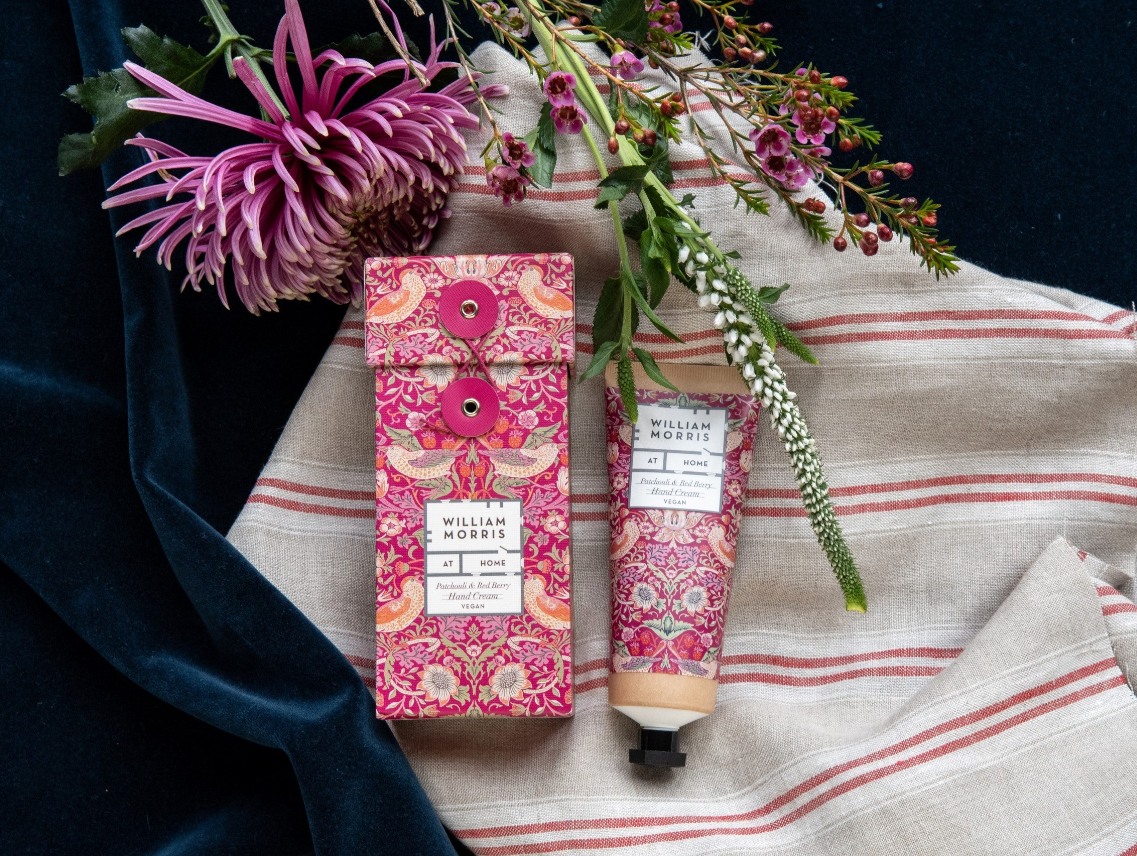 Das William Morris at Home 'Strawberry Thief' Handcreme 50 ml Set wird in einer passenden Geschenkbox geliefert. Es ist auf gestreiftem Tuch mit dunkelblauem Samt abgebildet und wird von lila und weißen Blumen akzentuiert.