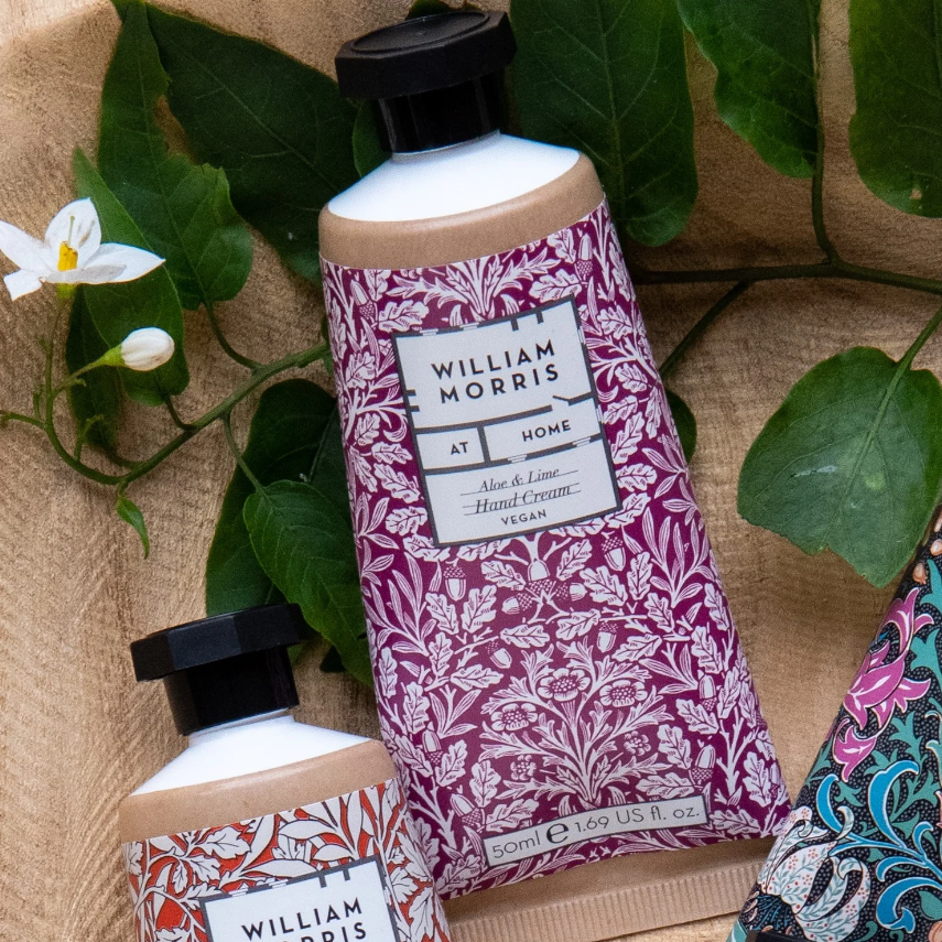 Eine Tube William Morris at Home 'Acorn' Handcreme 50 ml mit einem lila Blumenetikett ruht zwischen grünen Blättern und Blumen auf beigem Stoff.