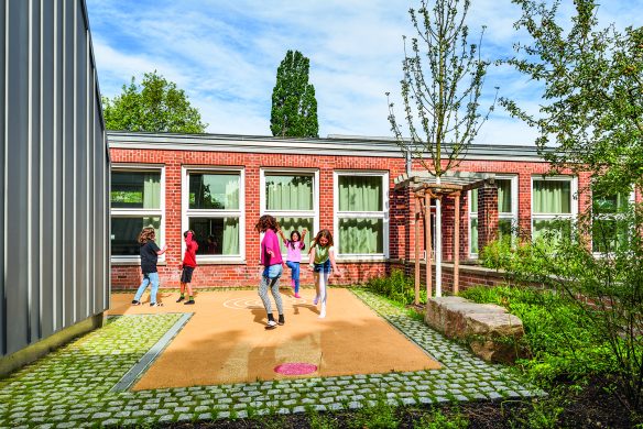 Kinder spielen gerne draußen auf einer gepflasterten Fläche neben einem modernen Backsteingebäude mit großen Fenstern und Grünflächen, das an einem sonnigen Tag das Ergebnis der durchdachten Gestaltung von Landschaftsarchitektur 2025 des Callwey Verlags zeigt.