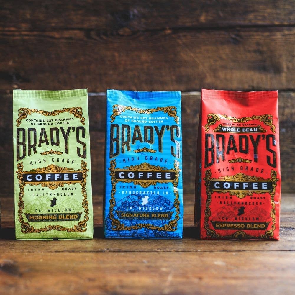 Drei farbenfrohe Tüten der Brady's Coffee Company - Morning Blend (grün), Signature Blend Kaffee (blau) und Espresso Blend (rot) - werden auf Holz präsentiert, jede mit einem kunstvollen Design und gefüllt mit erlesenen Kaffeespezialitäten.