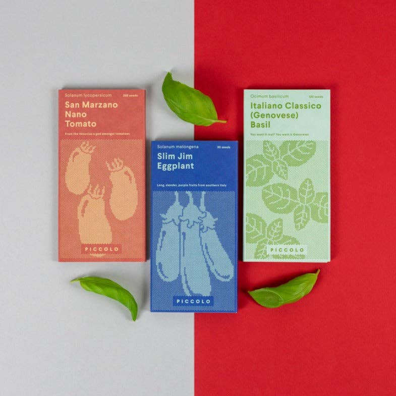 Drei leuchtende Piccolo Seeds-Päckchen - rot für San Marzano-Tomaten, blau für Aubergine Parmigiana und grün für Basilikum - sind auf einer grauen Fläche mit frischen Basilikumblättern angeordnet, ideal für Fans italienischer Kräutersamen.
