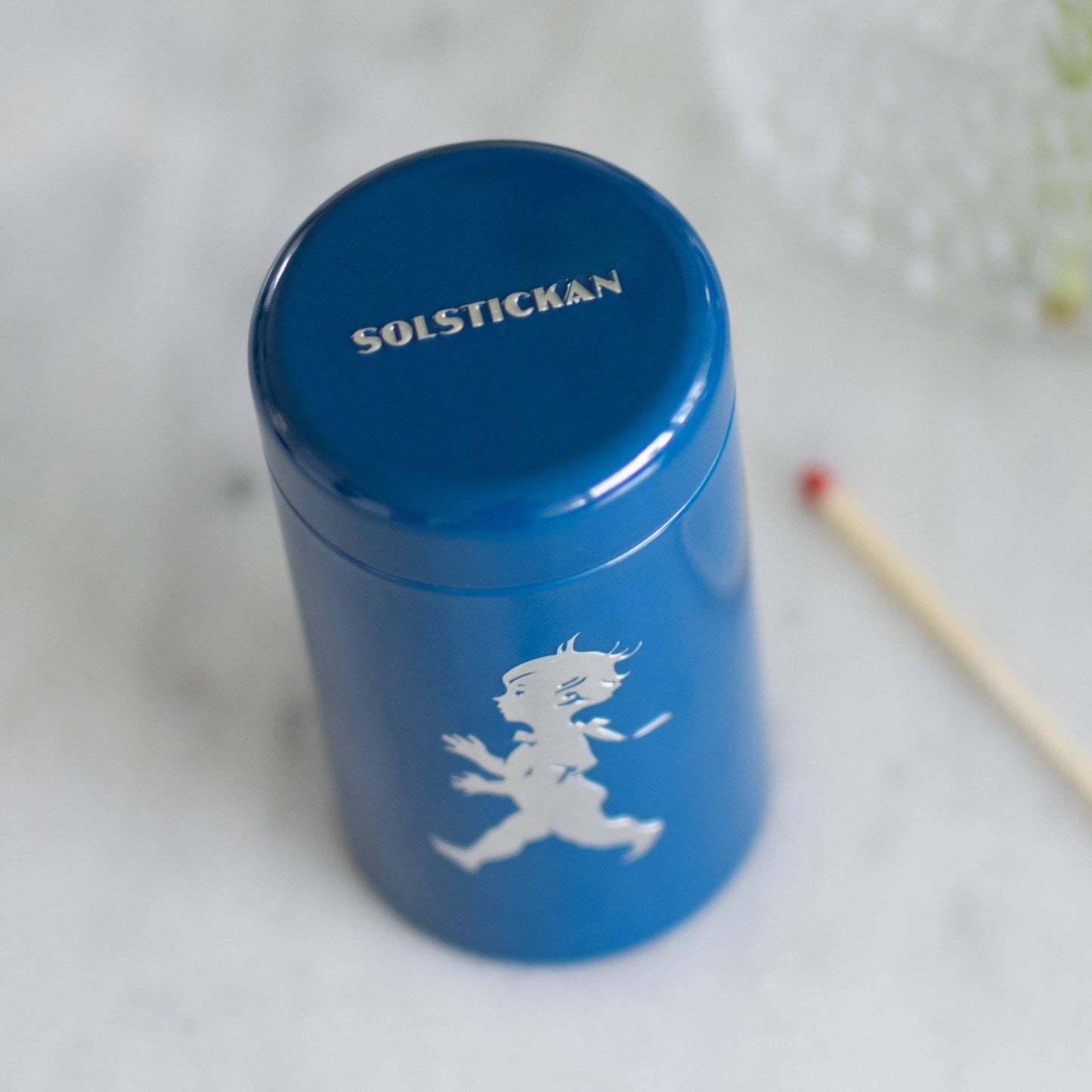 Der Solstickan Design - Passender Schlauch - Blau (nur EU) von Solstickan Design ist mit SOLSTICKAN auf dem Deckel und der kultigen Solsticke Boy-Silhouette versehen, die für echte schwedische Qualität steht. Er wird neben Streichhölzern auf einer weißen Fläche präsentiert.