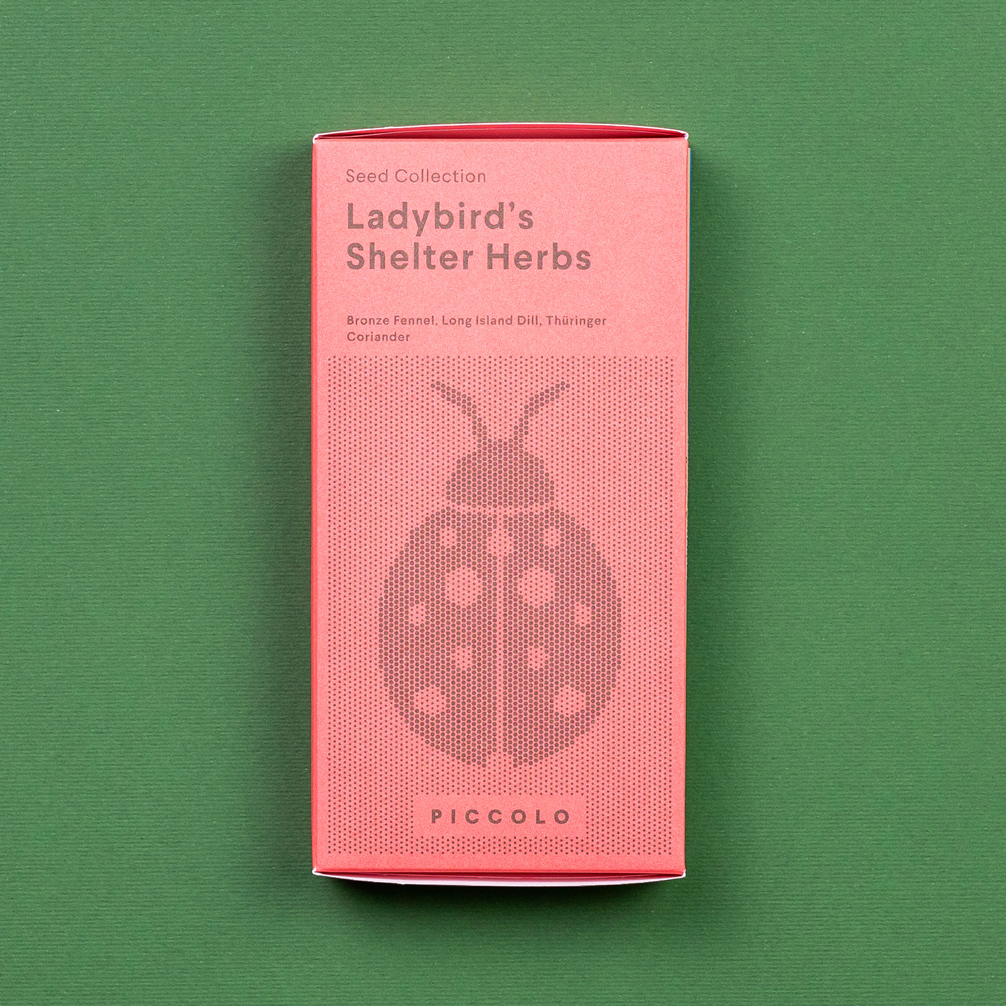 Eine rosafarbene Piccolo Seeds - Ladybird's Shelter Kräuter-Packung mit einer Marienkäfer-Illustration ruht auf Grün. Es hebt Samen hervor: Fenchel, Dill und Thaiingher Koriander der Marke Piccolo Seeds.