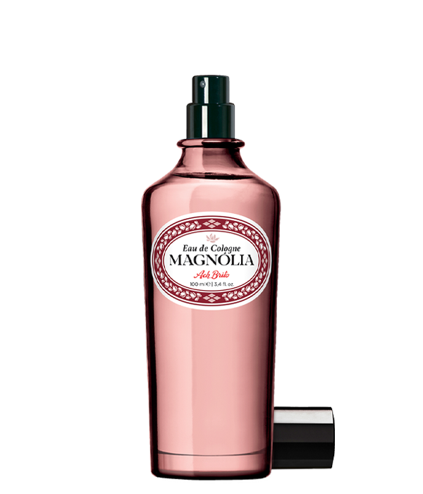 Ein rosafarbener Glasflakon von Ach.Brito Eau de Cologne Magnolia 100 ml mit dunklem Sprühkopf, weißem Etikett mit roter und schwarzer Schrift und daneben liegendem Verschluss, der den Duft andeutet.
