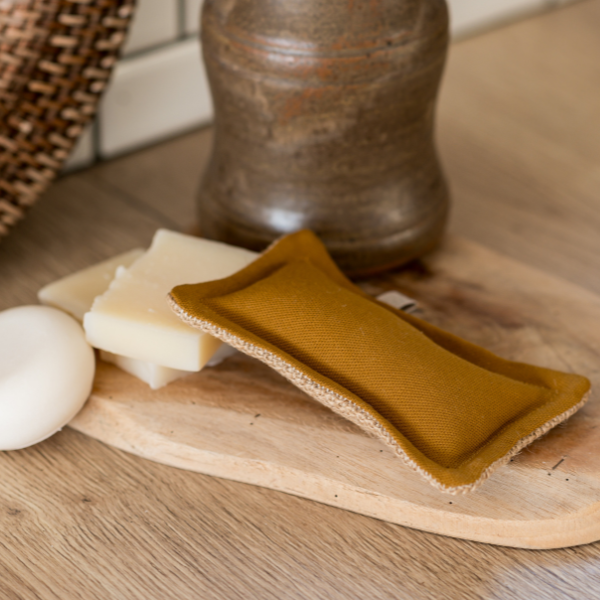 Der „Simple Things – Waschbarer Schwamm | Die Scharfen“ von Simple Things in Senfgelb aus Baumwolle/Jute liegt auf einem Holztablett neben zwei rechteckigen Seifen, einer runden weißen Seife und einem braunen Keramikbehälter.