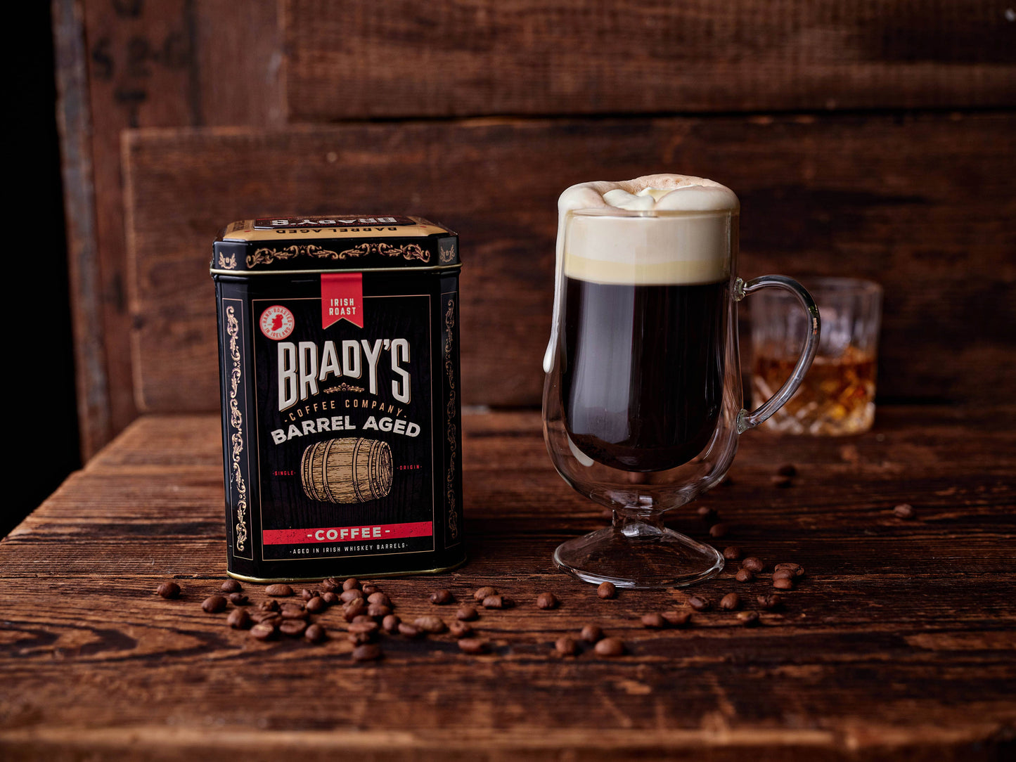 Ein Glas Irish Coffee mit Sahne steht neben der Brady's Coffee Company Kaffeedose mit ganzen Bohnen, im Fass gereift, irischer Whiskey; umgeben von Bohnen, mit einem Glas Whiskey im Hintergrund.