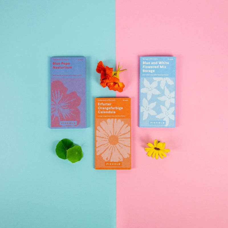 Drei Piccolo Seeds - Essbare Blumen Samenpakete mit floralen Designs liegen auf einem rosa-blauen Hintergrund, jeweils mit einer essbaren Blume - ideal als Blumen für Salate - daneben platziert.