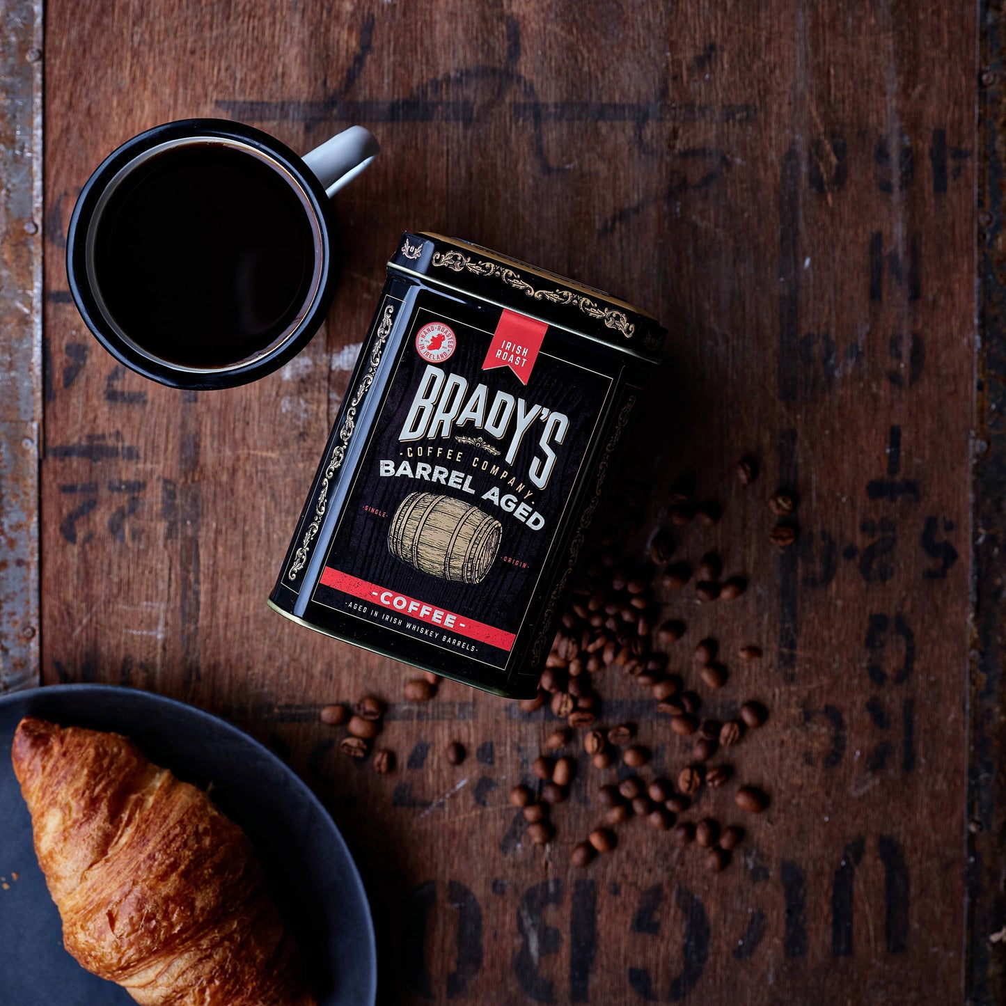 Eine Tasse schwarzen Kaffee, Brady's Coffee Company Kaffeedose mit ganzen Bohnen, im Fass gereift (irischer Whiskey), verstreute Bohnen und ein Croissant auf einem dunklen Teller stehen auf einer rustikalen Holzoberfläche.
