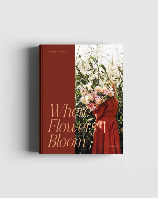 Das Buch „Where the Flowers Bloom“ von Cozy Publishing zeigt eine Person in einem orangefarbenen Kleid mit einem großen Blumenstrauß inmitten hoher Pflanzen, mit elegantem Goldtext auf rötlichem Einband. Perfekt für Blumenliebhaber.