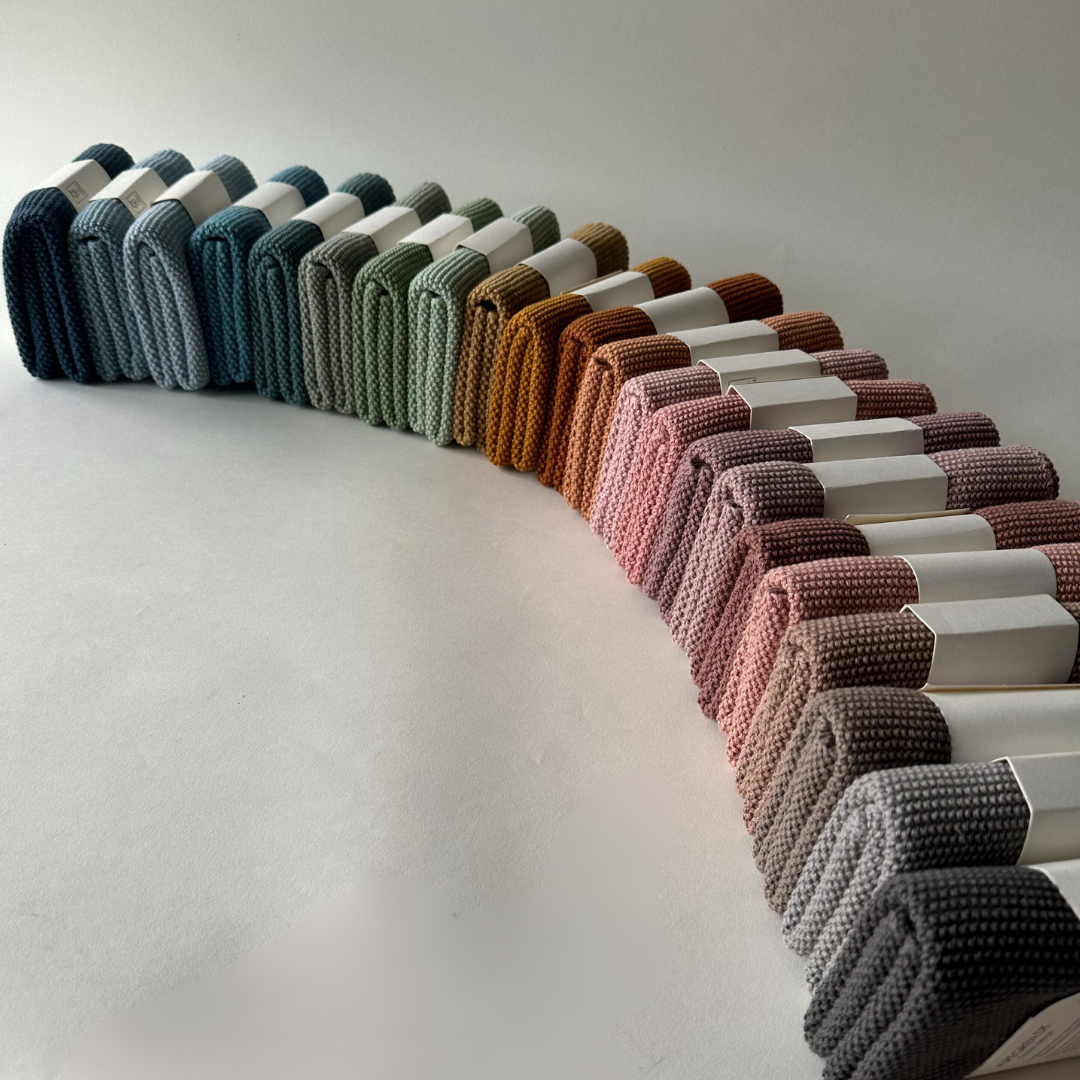 Eine geschwungene Reihe ordentlich gefalteter Küchentücher „Aspegren Denmark Solid“ in Pale Malve, jedes mit einem weißen Papierband umwickelt, auf einer weißen Oberfläche zwischen anderen bunten Baumwolltüchern angeordnet.