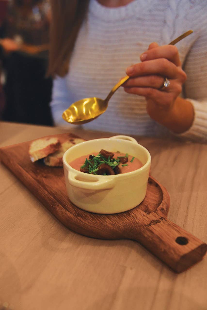 Eine Person im weißen Pullover hält einen goldenen Löffel über eine Schüssel Kräutersuppe, die neben Brotscheiben auf dem LE RÉGAL – Kleine Schaufel – Aperitifbrett aus französischem Eichenholz serviert wird.