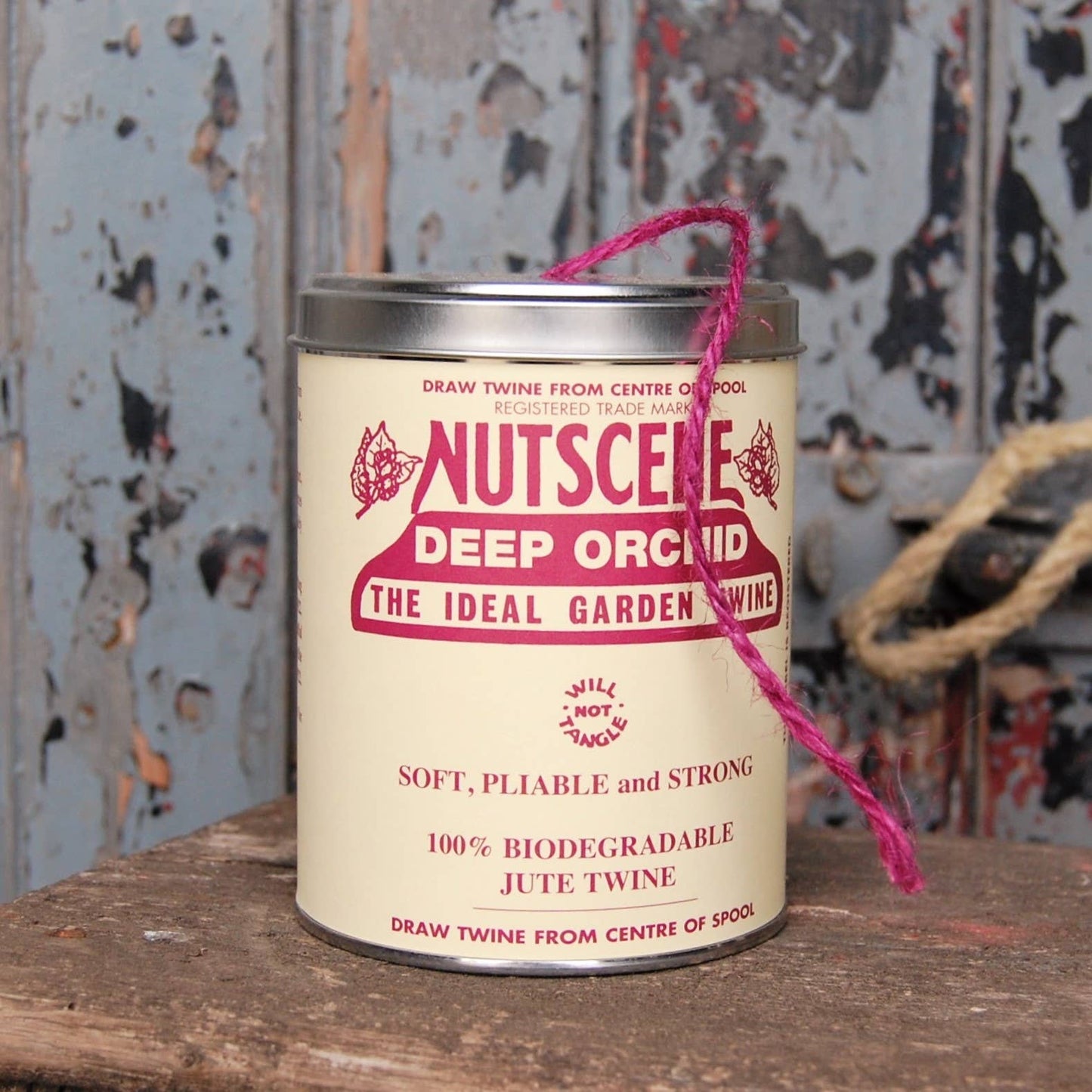 Eine ikonische Dose mit Nutscene Twine – Schnur in einer Dose (490 Fuß) in Deep Orchid von Nutscene Twines Ltd. steht auf einer rustikalen Holzoberfläche, die Schnur ragt aus der Oberseite heraus, mit verwitterter Farbe und Seil im Hintergrund.