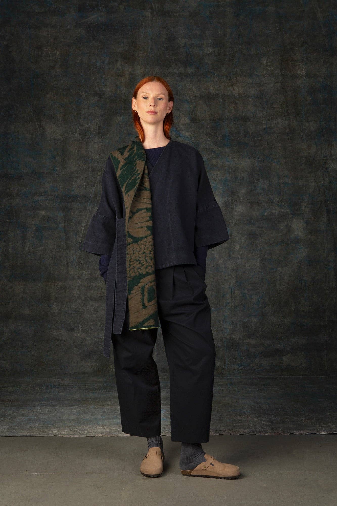Eine Person mit langen roten Haaren schaut nach vorne, trägt lockere dunkle Kleidung, beigefarbene Schuhe und einen Wolvis Schal aus Merinowolle - Furcula Wald Midi, der über eine Schulter drapiert ist, vor einem dunkel strukturierten Hintergrund.