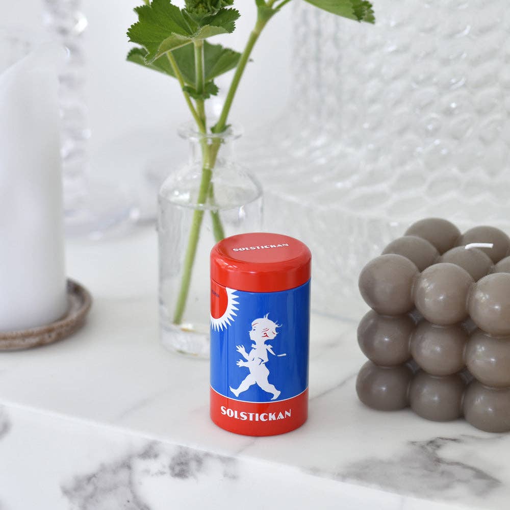 Ein Solstickan Design - Matches Tube - Originaldesign von Solstickan Design steht auf einer weißen Marmorfläche, umgeben von schwedischen Designelementen: eine graue Blasenkerze, eine Glasvase mit grünen Blättern und eine weiße Kerze.