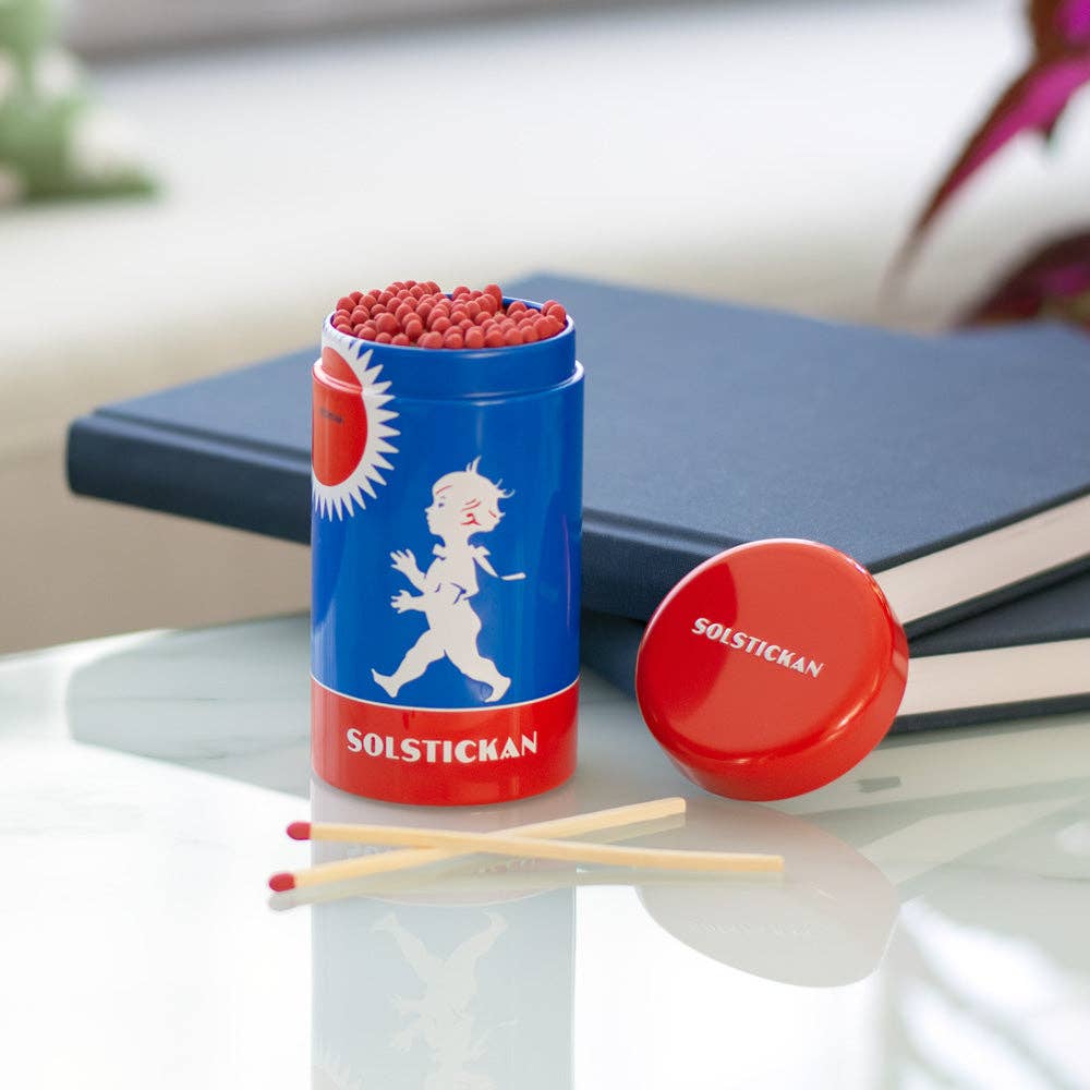 Eine Solstickan Design - Streichholzröhre - Originaldesign in Blau und Rot liegt auf einer weißen Fläche mit zwei Streichhölzern in der Nähe und geschlossenen Büchern dahinter. Die Röhre zeigt das ikonische schwedische Kind-und-Sonne-Design von Solstickan.