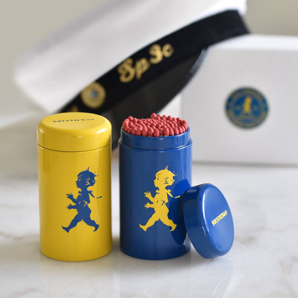 Zwei zylindrische Solstickan Design - Streichholzröhrchen in gelb und blau, jeweils mit dem Solsticke Boy. Die geöffnete blaue Röhre zeigt lange Streichhölzer mit roten Spitzen im Inneren. Der Hintergrund zeigt eine verschwommene weiße Schachtel und ein dunkles Band.