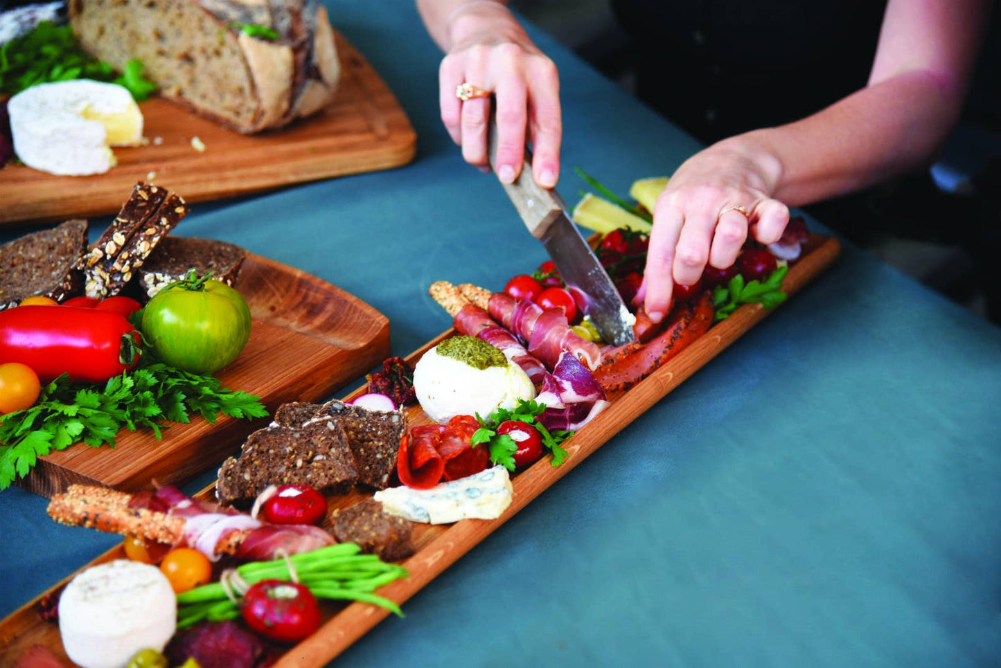 Eine Person schneidet Fleisch auf der LE RÉGAL - Lange Schaufel, einem großen Aperitifbrett aus Eichenholz von LE RÉGAL, umgeben von Käse, Wurst, Crackern, Kirschtomaten und Grünem; Ein weiteres Holzbrett mit Brot und Käse liegt auf einem blauen Tischtuch.
