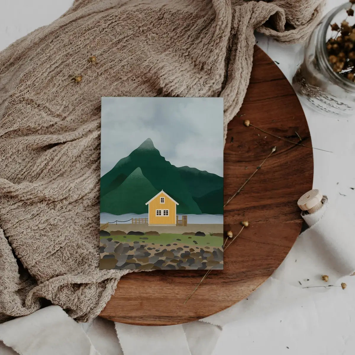 Die „Tilda&Theo – Postkarte – Norwegen Reise“ von Tilda&Theo zeigt die Illustration eines gelben Hauses und grüner Berge, dargestellt auf einer Holztafel mit einem beigen Tuch und einem Glas mit getrockneten Blumen.