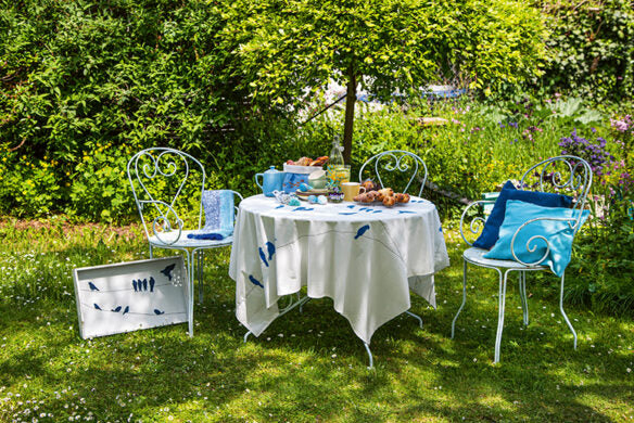 Die Tischdecke Frühlingstraum aus dem Callwey Verlag bringt Frühlingsflair in Ihren Teegarten: blaue Vögel auf weißem Stoff, Gebäck und Tassen stehen bereit, und blau gepolsterte Stühle umgeben von üppigem Grün - perfekt für Dekorationsideen.