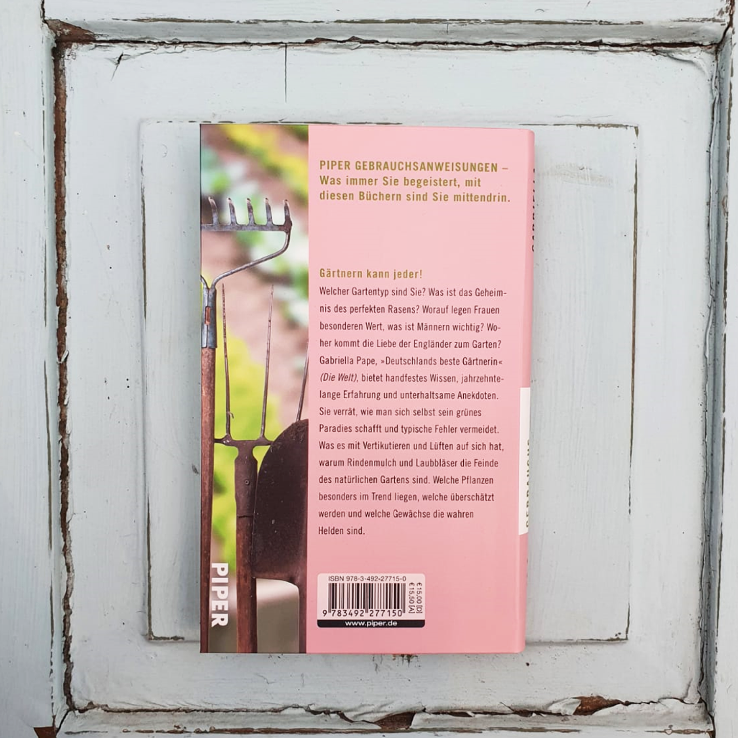 Die Rückseite von Gabriella Papes "Gebrauchsanweisung fürs Gärtnern | 7 Jahreszeiten, Praxis & Tipps Buch" ruht auf einer abgenutzten weißen Holzoberfläche, mit Gartengeräten, einer Pflanze auf der linken Seite und schwarzem Klappentext auf rosa Hintergrund.