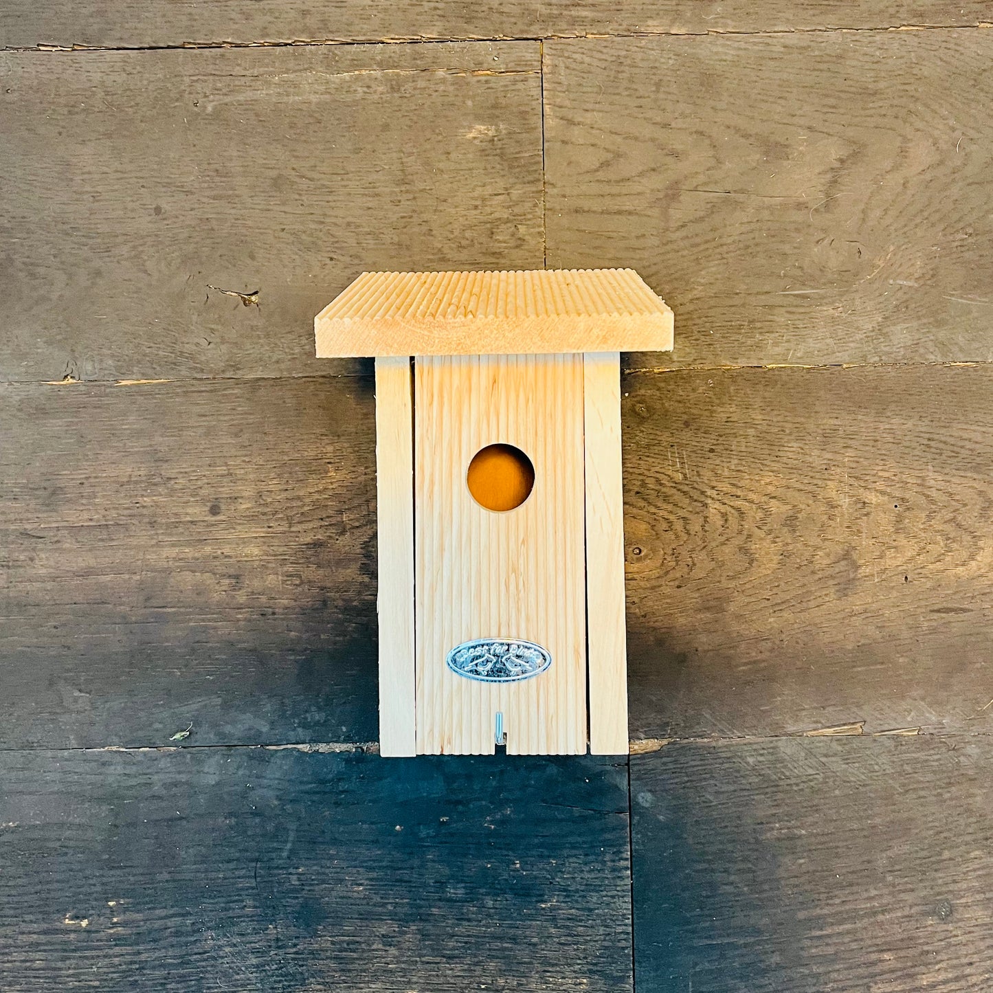 Der Esschert Nistkasten aus zertifiziertem Erlenholz verfügt über einen kleinen runden Eingang, ein flaches, strukturiertes Dach und ein dekoratives ovales Emblem. An einer Holzwand montiert ist es ideal für verschiedene Vogelarten.