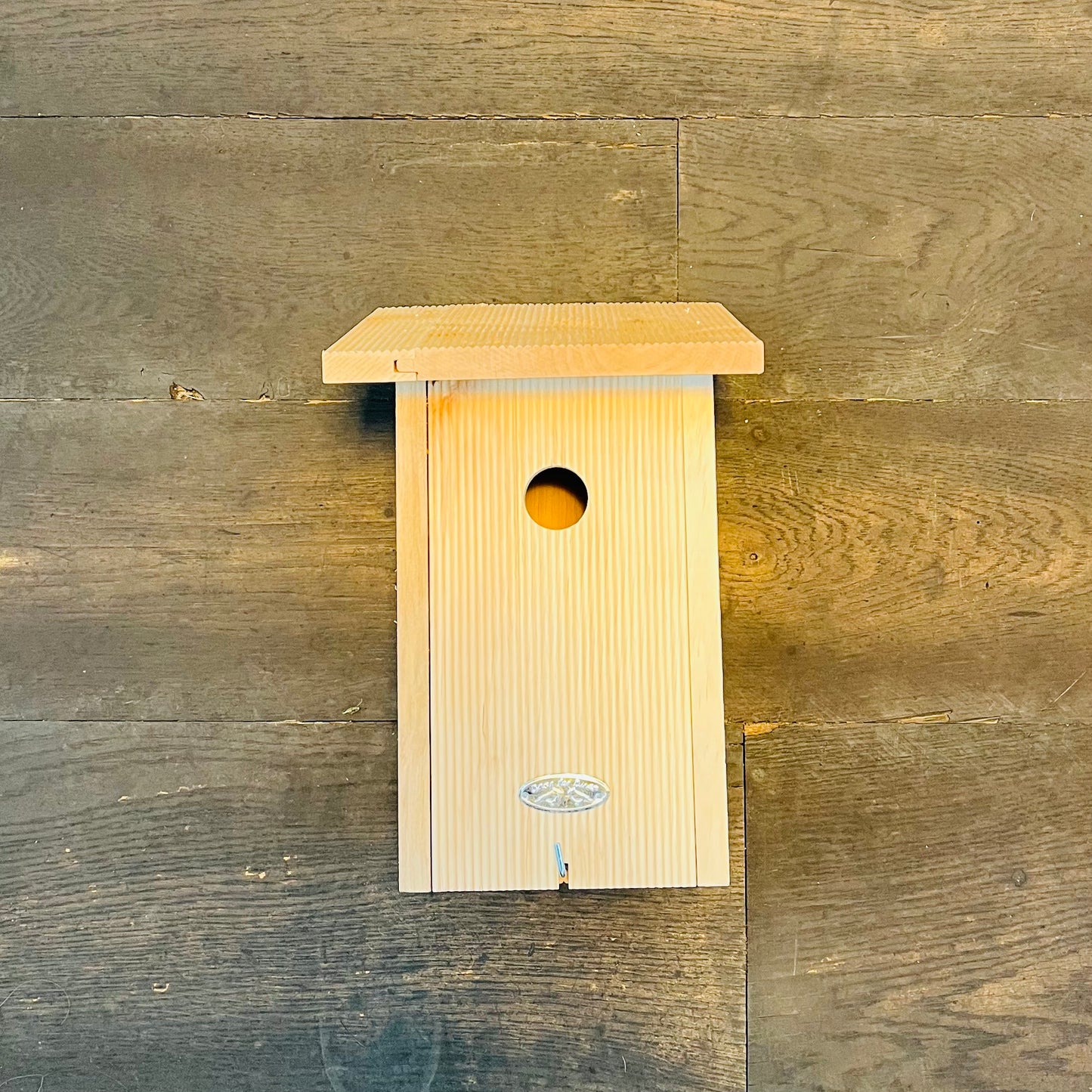 Der Esschert Nistkasten aus zertifiziertem Erlenholz mit schrägem Dach und runder Öffnung wird an einer rustikalen Holzwand montiert. Ein kleines Metallemblem am Sockel verleiht ihm Charme und macht ihn ideal zum Anlocken verschiedener Vogelarten.