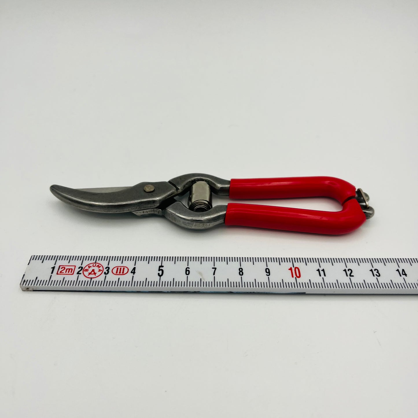 Ein Paar Esschert Mini Obst- und Blumenscheren mit roten Griffen und scharfen Klingen liegt neben einem Maßband auf einer weißen Fläche. Die Scheren, ideal zum Schneiden von Blumen, sind etwa 18 cm lang.