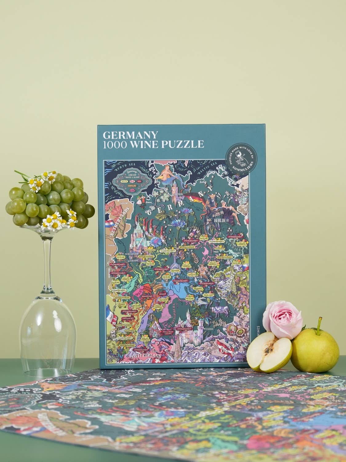 Eine farbenfrohe Water & Wines - Wein Puzzle - Deutschland Box steht aufrecht auf einem Tisch neben einem Glas mit grünen Trauben, einer rosa Rose und einem grünen Apfel, wobei ein Teil des zusammengesetzten Puzzles deutsche Weinregionen im Vordergrund zeigt.