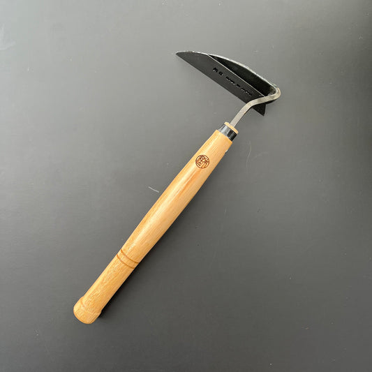 Die Niwaki-Unkrauthacke von Niwaki mit ergonomischem Design, gebogener schwarzer Metallklinge und langem hellem Holzgriff liegt auf einer dunklen Oberfläche – perfekt zum Entfernen von Unkraut in Ihrem Garten.