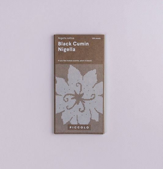 Eine Packung Piccolo Seeds – Nigella Schwarzkümmel liegt auf einer helllavendelfarbenen Oberfläche und zeigt ein weißes Blumenmuster sowie einen Text, der darauf hinweist, dass sie 100 Samen Nigella sativa der Marke Piccolo Seeds enthält.