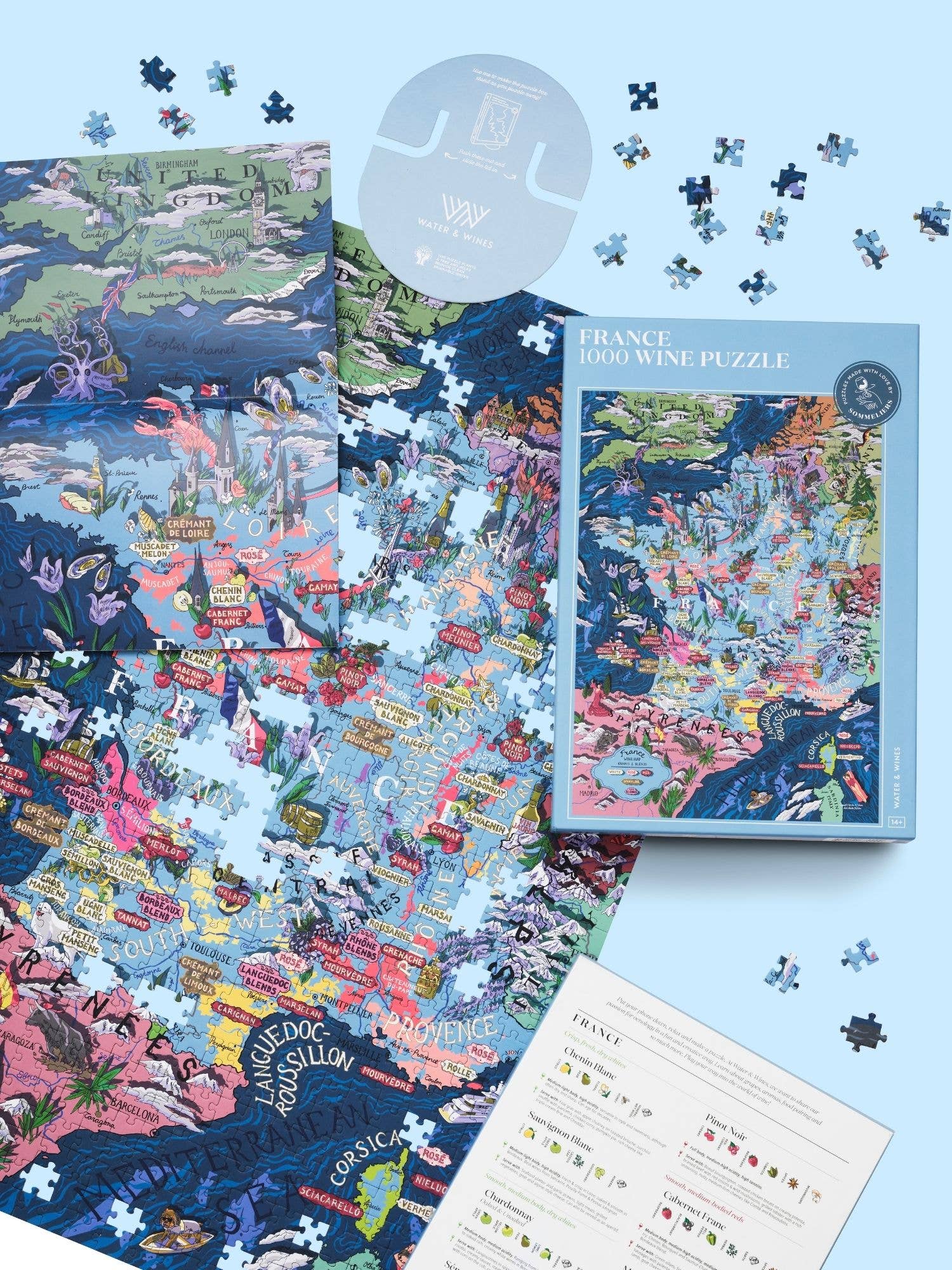 Ein teilweise zusammengesetztes Water & Wines - Wein Puzzle - Frankreich von Water & Wines liegt auf einer hellblauen Fläche mit losen Teilen drum herum. Die Puzzlebox, das Referenzblatt und eine runde blaue Karte liegen ebenfalls in der Nähe.