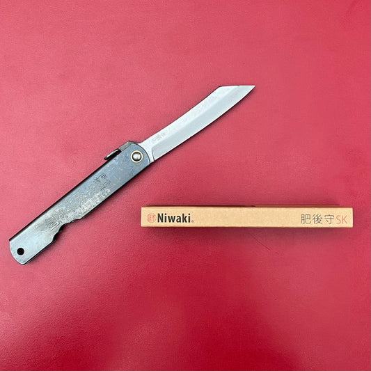 Ein Niwaki SK Higonokami-Klappmesser mit silbernem Griff und scharfer Klinge liegt teilweise geöffnet neben seiner braunen Niwaki-Box auf einer roten Oberfläche.