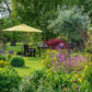 Ein üppiger Garten mit leuchtenden "Stauden im Garten" aus dem Callwey Verlag und grünen Sträuchern umgibt einen Holztisch und Stühle unter einem großen gelben Sonnenschirm auf einem grasbewachsenen Rasen mit Bäumen und Sträuchern im Hintergrund.