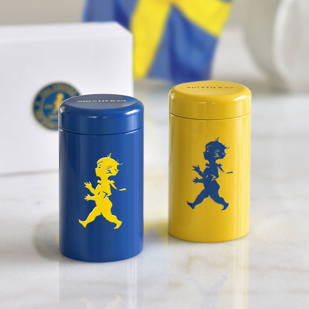 Die Solstickan Design - Match Tube - Gelb/Blau von Solstickan Design, mit dem klassischen Solsticke Boy, steht auf Marmor. Eine blaue und eine gelbe Dose erinnern an schwedische Qualität und fassen extralange Streichhölzer (nur EU).