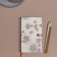 Der Tinne Mia Notizbuchknopf Leinen "Amour des lilas" FSC Mix liegt neben zwei Bleistiften und einem Kugelschreiber auf einer rosa-braunen Fläche, wobei in der linken oberen Ecke ein Teller und eine Tasse zu sehen sind.