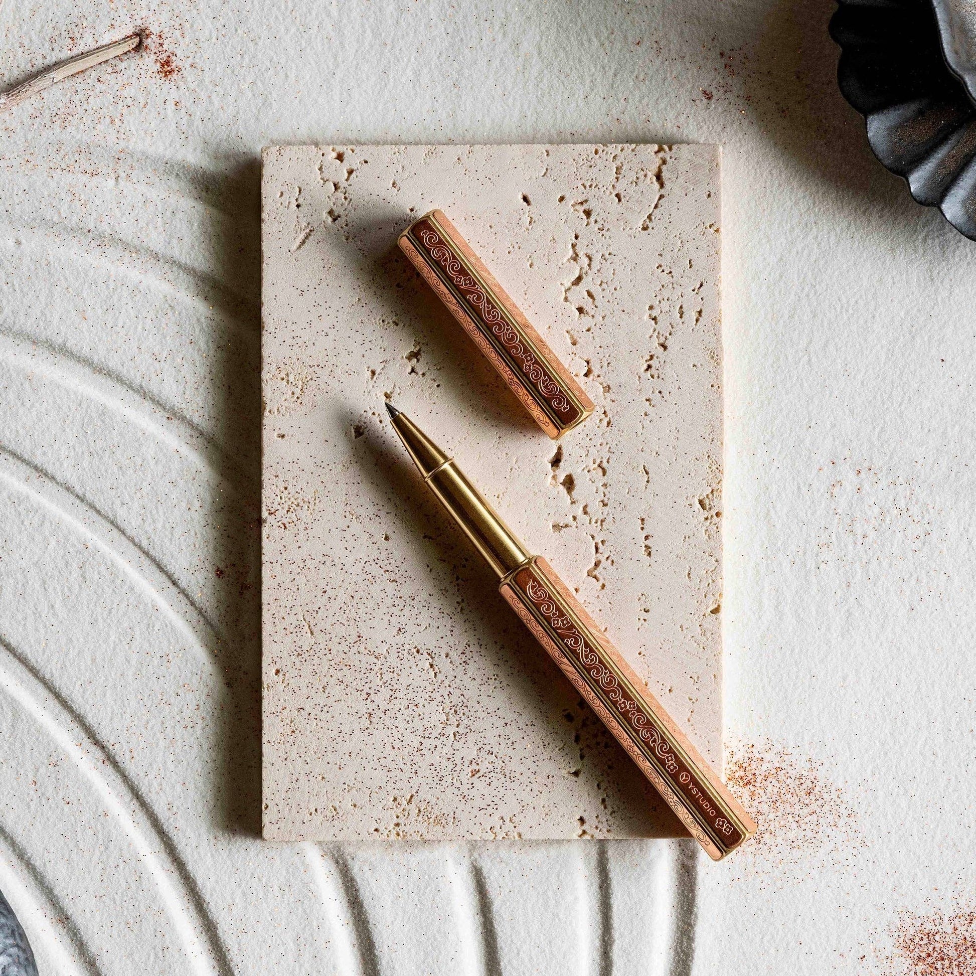 Der ENTREPÔT YSTUDIO Klassische Renaissance KAZARI KANAGU ORCHID Tintenroller, ein dekorativer Stift aus Gold und Kupfer, ruht auf einer strukturierten beigen Steinplatte auf weißem Grund mit kreisförmigen Rillen und Pulver.