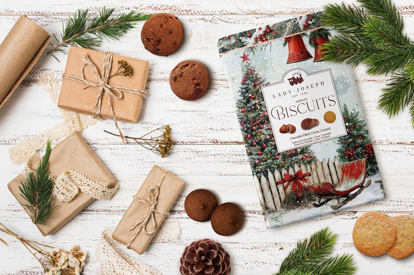 Ein festliches Flat Lay zeigt die Lady Joseph - Sortiment von Keksen in einer Metallgeschenkbox für Weihnachten, eingepackte Geschenke, Tannenzapfen, Grünzeug und gemischte Kekse auf einer weißen Holzoberfläche.